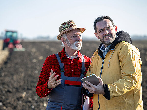 Ein Landwirt und ein Mann mit Tablet besprechen sich auf einem Feld. Dabei befindet sich im Hintergrund ein Traktor. 