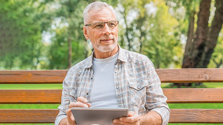Ein älterer Mann sitzt auf einer Parkbank und hat ein Tablet in der Hand.