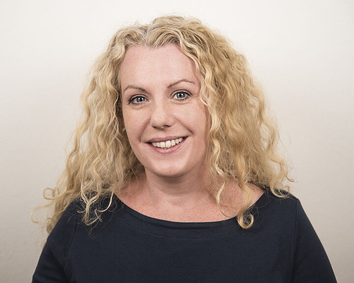 Porträtfoto einer Frau mit blonden Locken, die ein schwarzes Oberteil trägt und lächelt.
