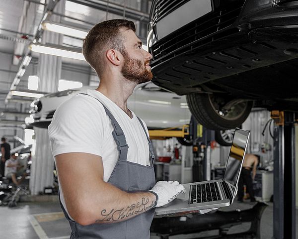 Ein Mechaniker steht mit Laptop vor einer Autohebebühne und sieht sich das Auto in der Werkstatt an.