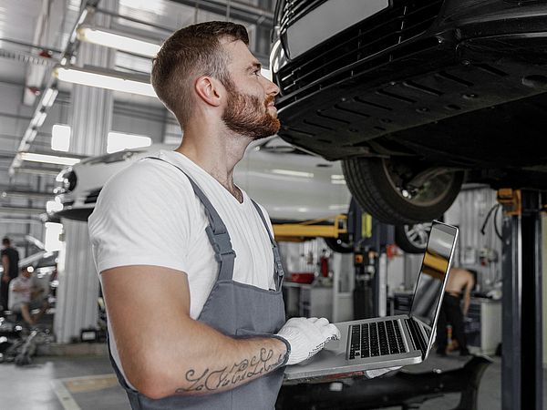 Ein Mechaniker steht mit Laptop vor einer Autohebebühne und sieht sich das Auto in der Werkstatt an.