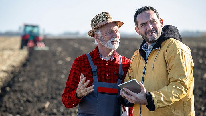 Ein Landwirt und ein Mann mit Tablet besprechen sich auf einem Feld. Dabei befindet sich im Hintergrund ein Traktor. 