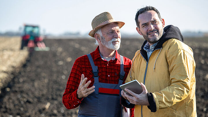 Ein Landwirt und ein Mann mit Tablet besprechen sich auf einem Feld. Dabei befindet sich im Hintergrund ein Traktor. 