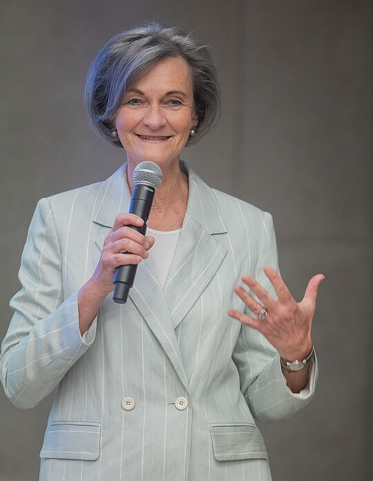 Porträt von Finanzexpertin Beatrice Schobesberger in einem hellen Blazer mit Nadelstreifen, die lächelnd ein Mikrofon hält und spricht.