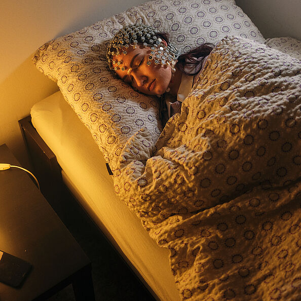 Eine Person schläft mit einer EEG-Haube auf dem Kopf. Sie hat vergessen, die Nachttischlampe auszuschalten.
