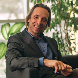 Ein Mann mit schulterlangen braunen Haaren sitzt in einem Sessel und lacht in die Kamera. Seine Finger sind ineinander verschränkt, seine Augen halb geschlossen. 