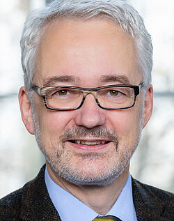 Porträtbild von einem Mann mit weißen Haaren und etwas Bart im Anzug mit Krawatte