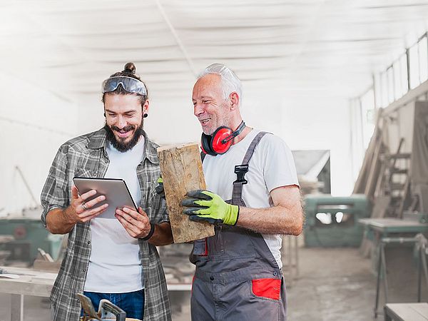 Ein älterer Mann mit einem Stück Holz in der Hand und ein junger Mann blicken auf ein Tablet einer Werkstatt.