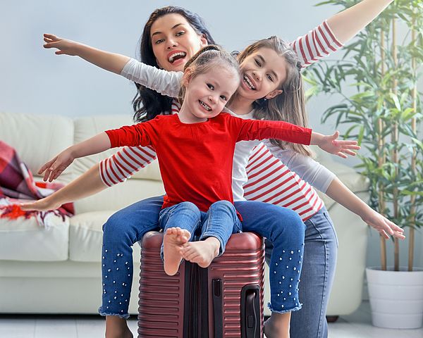Eine glückliche Familie bereitet sich auf eine Reise vor. Die zwei Töchter sitzen auf dem gepackten Koffer und strecken die Arme in die Luft.