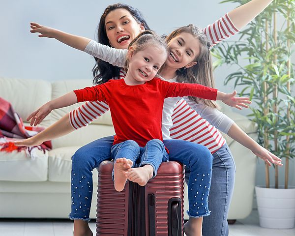 Eine glückliche Familie bereitet sich auf eine Reise vor. Die zwei Töchter sitzen auf dem gepackten Koffer und strecken die Arme in die Luft.