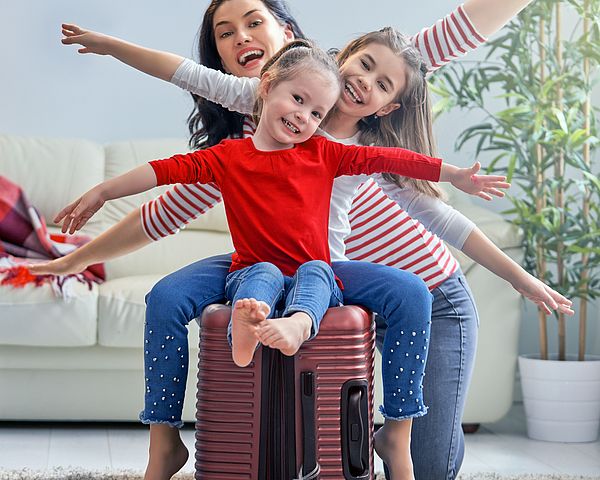 Glückliche Familie bereitet sich auf eine Reise vor. Die zwei Töchter sitzen auf dem gepackten Koffer und strecken die Arme in die Luft.