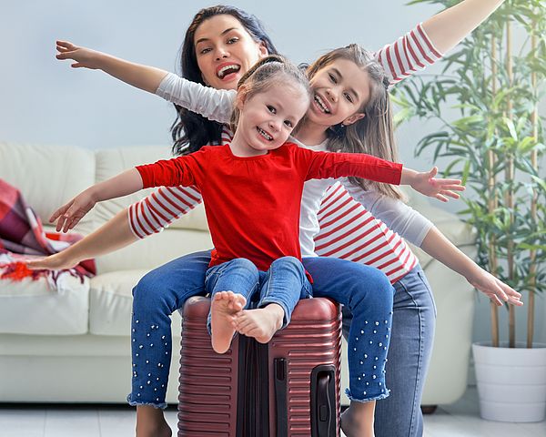 Eine glückliche Familie bereitet sich auf eine Reise vor. Die zwei Töchter sitzen auf dem gepackten Koffer und strecken die Arme in die Luft.