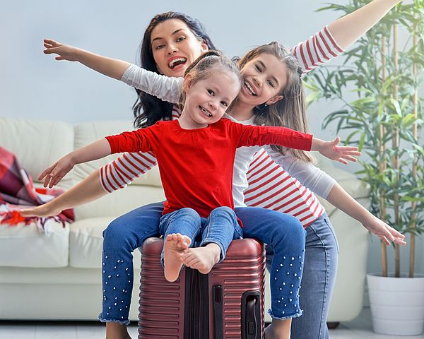 Eine glückliche Familie bereitet sich auf eine Reise vor. Die zwei Töchter sitzen auf dem gepackten Koffer und strecken die Arme in die Luft.