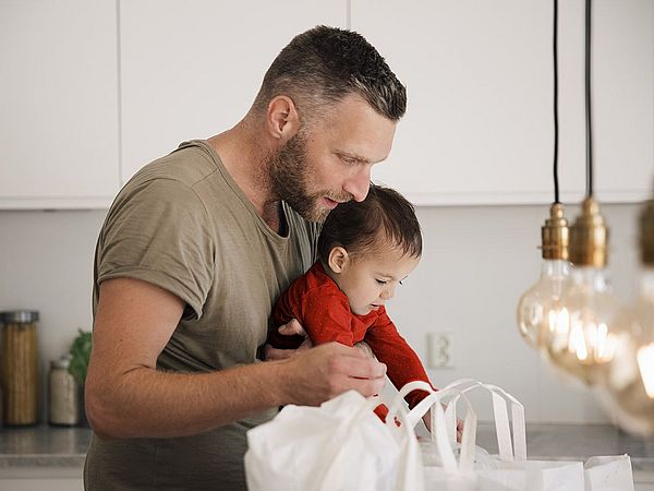 Ein Vater hält sein Kind im Arm. Dabei schauen beide voller Neugier in eine Tasche 