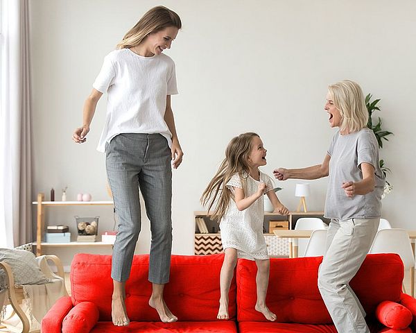 Ein glückliches junges Mädchen springt mit ihrer Mutter und ihrer Großmutter auf dem Sofa. 