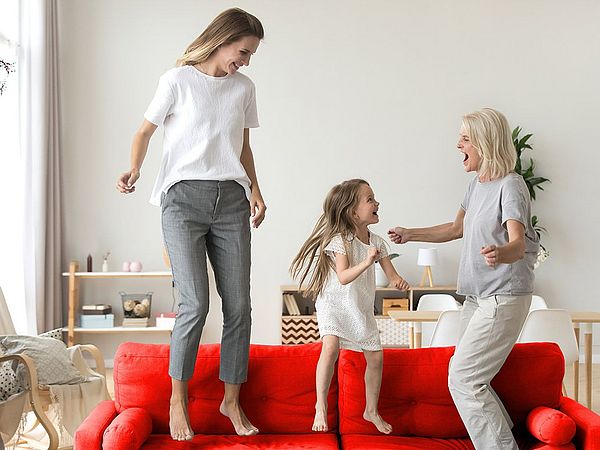 Ein glückliches junges Mädchen springt mit ihrer Mutter und ihrer Großmutter auf dem Sofa. 