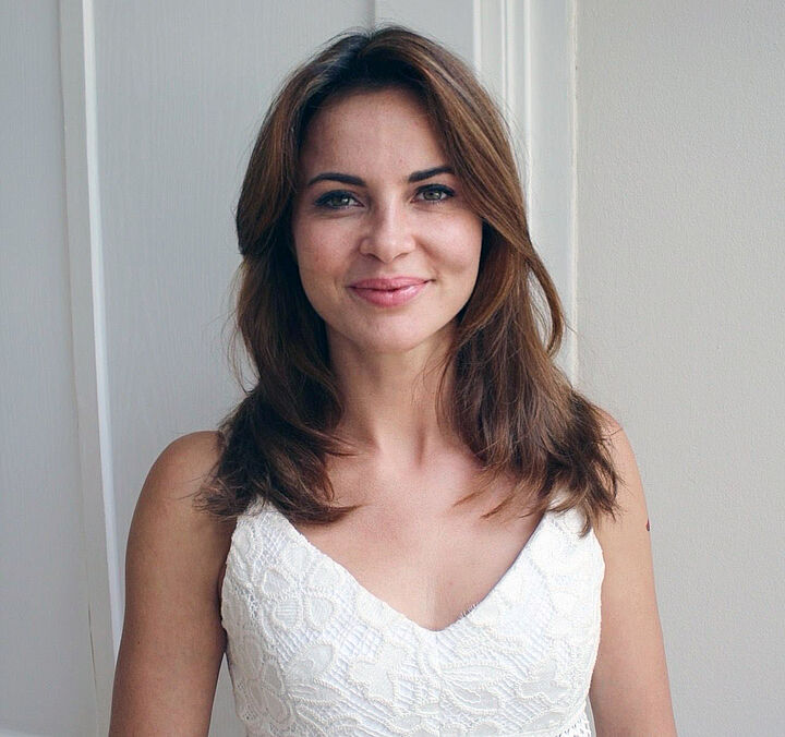 Portrait einer lächelnden Frau mit schulterlangen, braunen Haaren in einem weißen, spitzenbesetzten Oberteil vor hellem Hintergrund.
