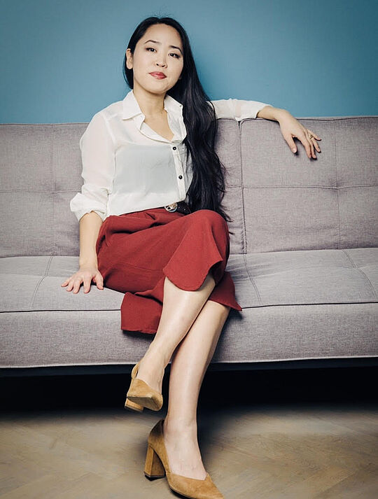 Psychotherapeutin Hiina Smyth sitzt entspannt auf einer grauen Couch, trägt eine weiße Bluse, eine rote Hose und helle Pumps.