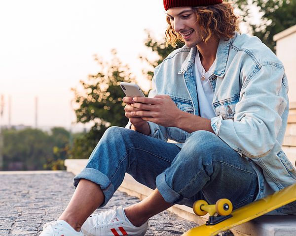 Ein junger Skateboard-Fahrer sitzt auf Stufen im Freien und tippt auf seinem Smartphone.