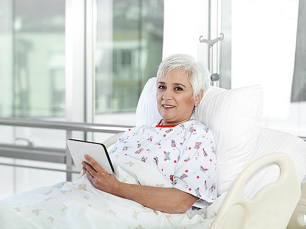 Eine ältere Dame befindet sich im Krankenbett und hält dabei lächelnd ein Tablet in der Hand.