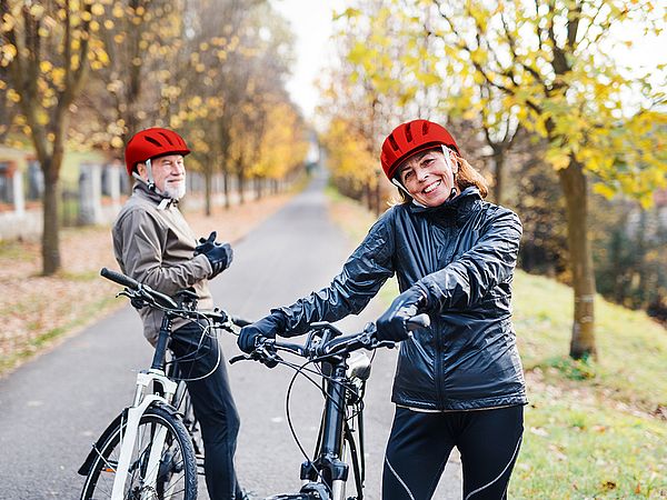Ein glückliches Seniorenpaar in Sportbekleidung steht mit Fahrrädern auf einer Straße in der Natur.