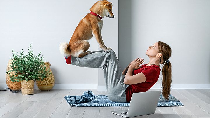 Eine junge Frau macht Sit-ups auf einer Gymnastikmatte in ihrer Wohnung, während ihr Hund auf ihren Beinen sitzt.