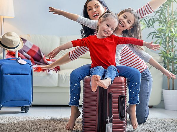 Eine glückliche Familie bereitet sich auf eine Reise vor. Die zwei Töchter sitzen auf dem gepackten Koffer und strecken die Arme in die Luft.