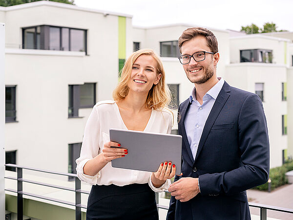 Eine Geschäftsfrau und ein Geschäftsmann befinden sich auf einer Dachterrasse. Dabei lächeln beide in die Kamera und halten ein Tablet in der Hand. 