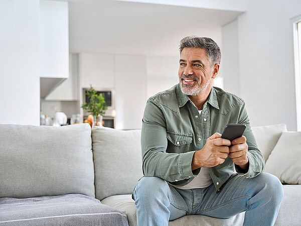 Mann mittleren Alters sitzt auf einem Sofa in einem hellen Wohnzimmer, hält ein Smartphone und lächelt leicht zur Seite.
