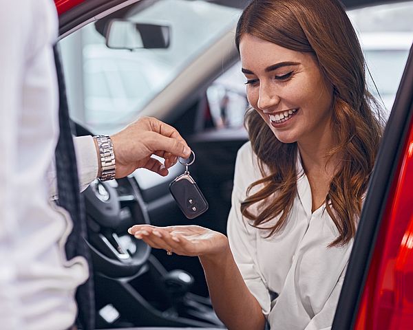 Glückliche junge Frau sitzt in ihrem neuen Auto und nimmt den Autoschlüssel vom Verkäufer entgegen.