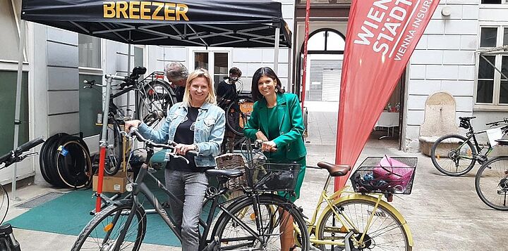 Zwei lächelnde Frauen mit Fahrrädern vor einem Fahrradservice-Stand.