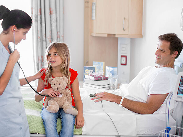 Krankenhausszene in einem hellen Patientenzimmer: Eine Pflegekraft hört mit einem Stethoskop das Herz eines Mädchens ab, das auf dem Bett sitzt und einen Teddybären im Arm hält. Daneben liegt ein Mann im Krankenhausbett und lächelt, angeschlossen an medizinische Geräte.