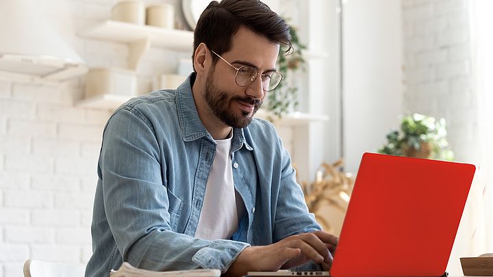 Ein junger, dunkelhaariger Mann mit Bart und Brille sitzt am Schreibtisch und arbeitet am Laptop.