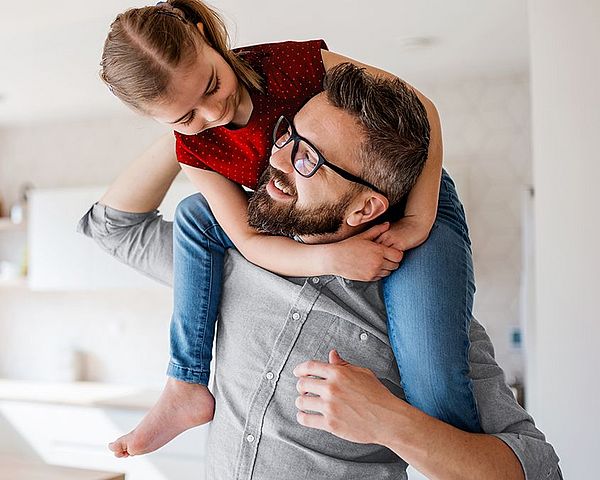 Ein Mann mit Brille trägt zu Hause seine Tochter auf den Schultern und sehen sich dabei direkt an 