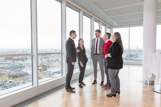 Mitarbeiterinnen und Mitarbeiter der Wiener Städtischen stehen in einem Raum mit großen Fenstern beisammen und unterhalten sich.