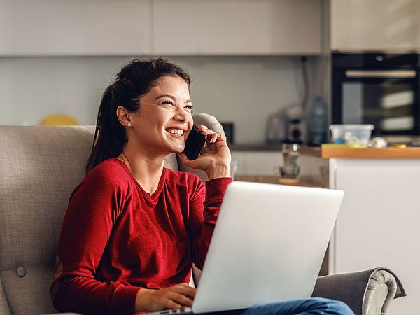 Eine glückliche Frau sitzt mit ihrem Laptop auf einem Sofa im Wohnzimmer und telefoniert