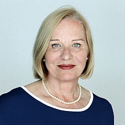 Eine Frau mit kinnlangen blonden Haaren trägt ein dunkles Oberteil mit weißem Rand und eine Perlenkette.