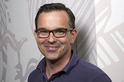 Ein Mann trägt eine schwarze Brille und ein dunkles Polo-Shirt und lehnt an einer gemusterten Tapetenwand.