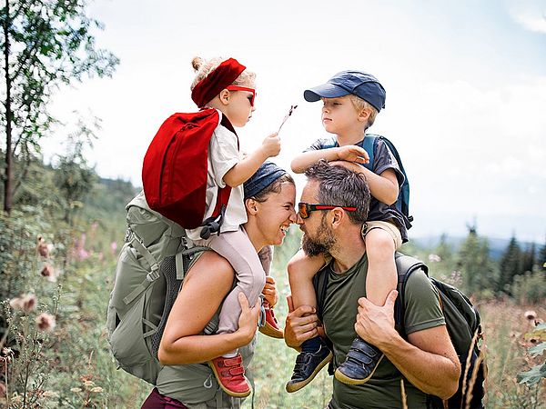 Eine junge glückliche Familie mit zwei Kindern wandert in den Bergen. Die Kinder werden auf den Schultern getragen. 