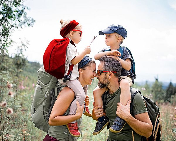 Eine junge glückliche Familie mit zwei Kindern wandert in den Bergen. Die Kinder werden auf den Schultern getragen. 