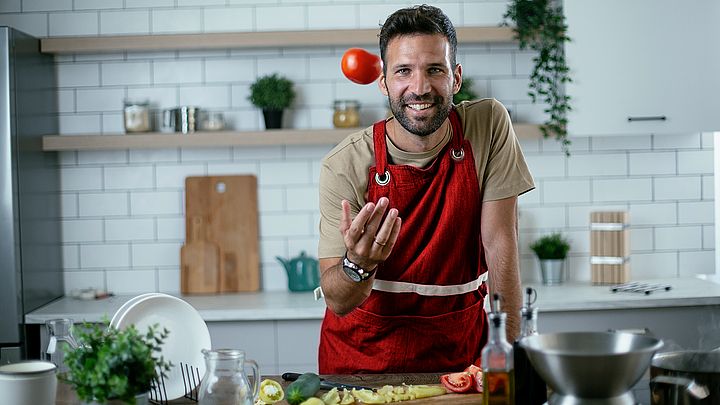 Ein junger Mann bereitet in der Küche das Gemüse zu und wirft eine Tomate in die Höhe.