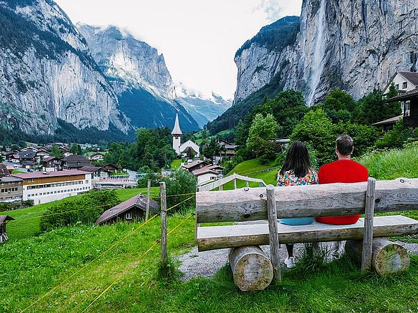 Ein Paar sitzt auf einer Bank in der Natur und blickt auf ein Dorf, welches umgeben von Bergen ist.