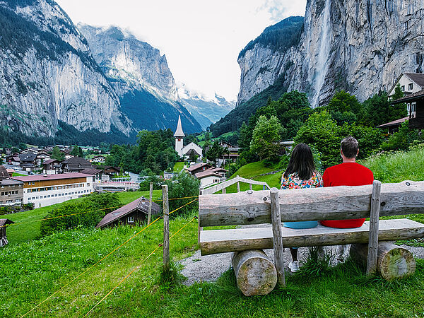 Ein Paar sitzt auf einer Bank in der Natur und blickt auf ein Dorf, welches umgeben von Bergen ist.