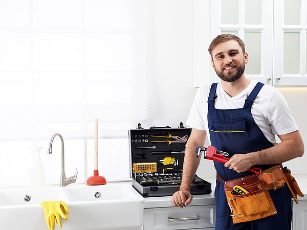Handwerker steht mit Werkzeug und Arbeitsgewand vor einem Küchenkasten.