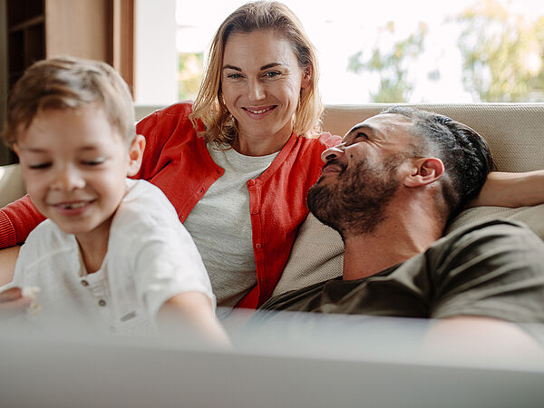 Familiäre Szene im Wohnzimmer: Eine Frau und ein Mann sitzen entspannt auf einem Sofa und lächeln sich an, während ein kleines Kind im Vordergrund spielt und lachend in Bewegung ist.