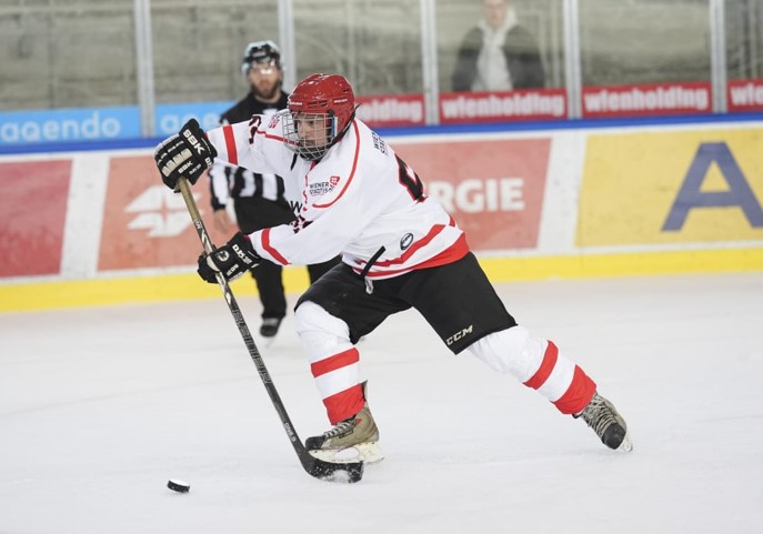 Eine Person in Eishockeyausrüstung bewegt sich mit mit Schläger und Puck über das Spielfeld, im Hintergrund steht ein Schiedsrichter.