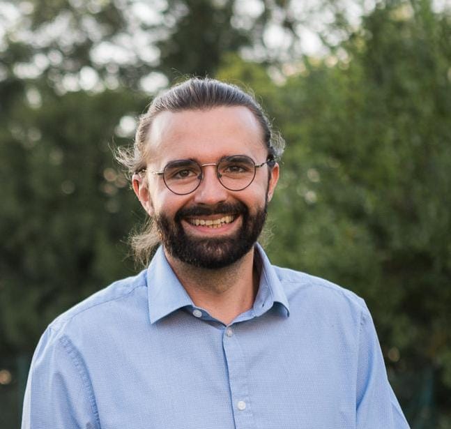 Portraitfoto eines lachenden Mannes mit dunklen Haaren, Zopf, Bart und Brille.