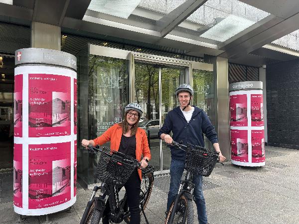 Zwei Personen (Frau und Mann) auf einem E-Bike sitzend vor dem Bürogebäude.